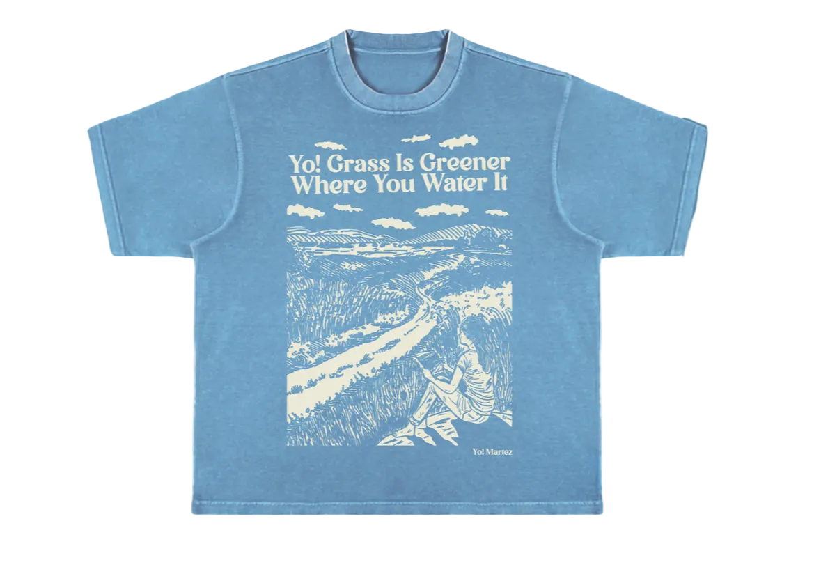 A blue crewneck t-shirt with a graphic print of a grassy field and the phrase 'Yol grass is greener where you water it'.