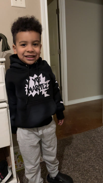 A young boy smiling while wearing a black pullover hoodie with a white comic-style graphic that says 'YOU MARTEZ' and light gray sweatpants, standing indoors.