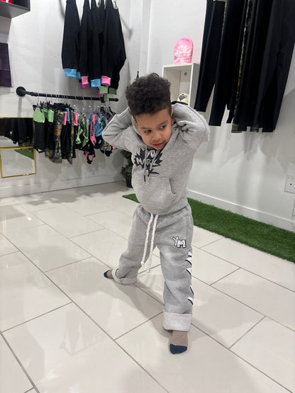 A young boy wearing a light gray tracksuit with black graphic details on the hoodie and white stripes on the pants, posing in a clothing store.