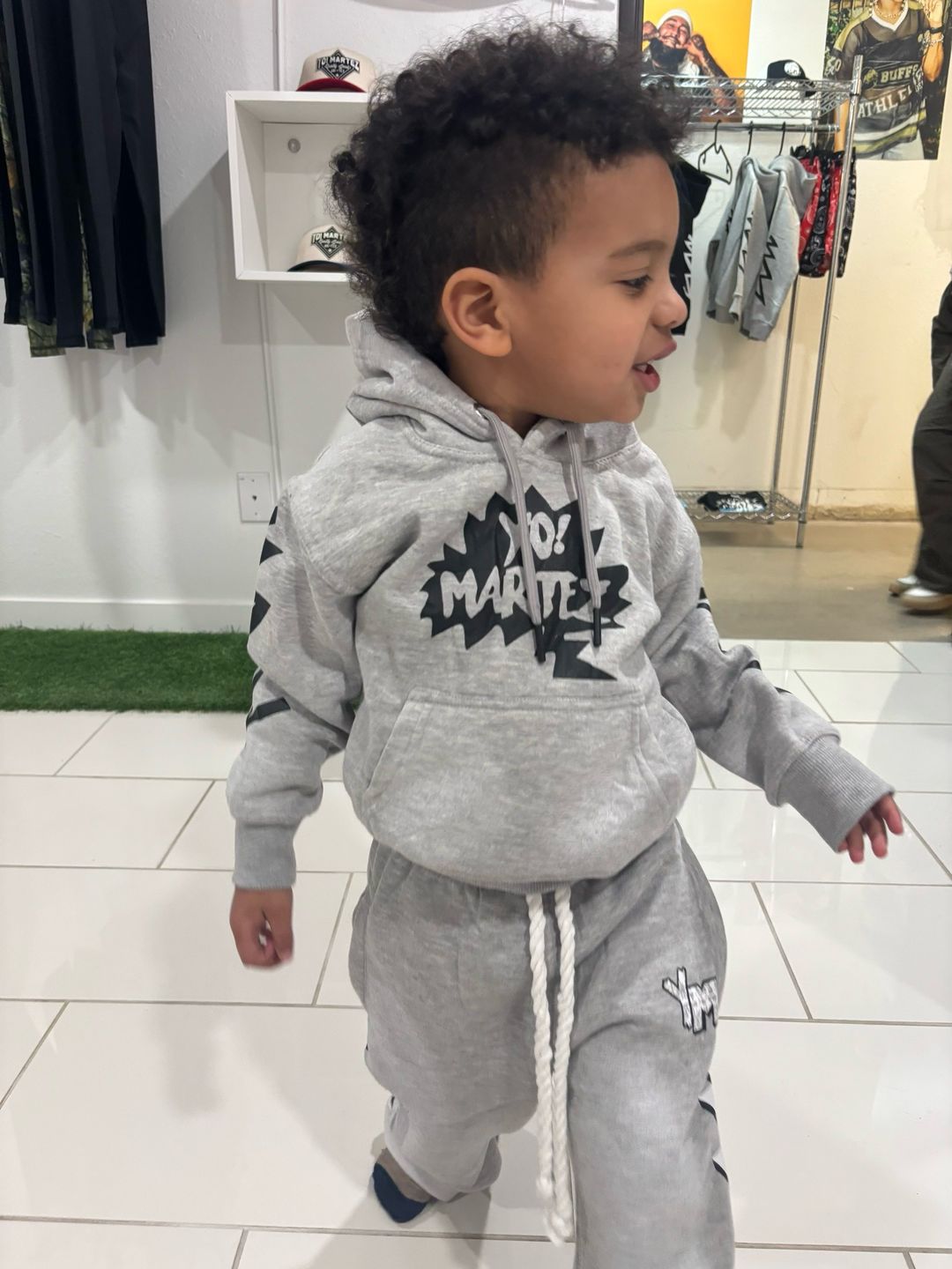 A toddler wearing a gray heathered hoodie and matching sweatpants set with black and white graphics, standing in a clothing store.