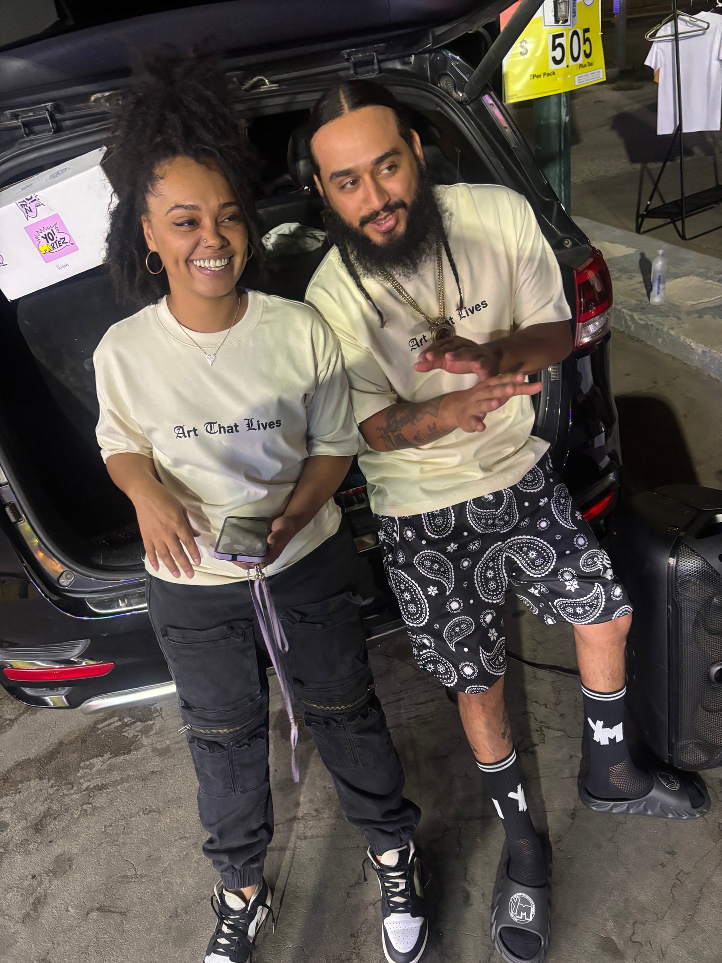 Crystal and Angel lying inside the open trunk of a black car, wearing matching cream-colored t-shirts with 'Art That Lives' printed on the front, dark jeans, and black socks with white logos. The photo is taken in a casual, selfie-style orientation on a concrete floor.