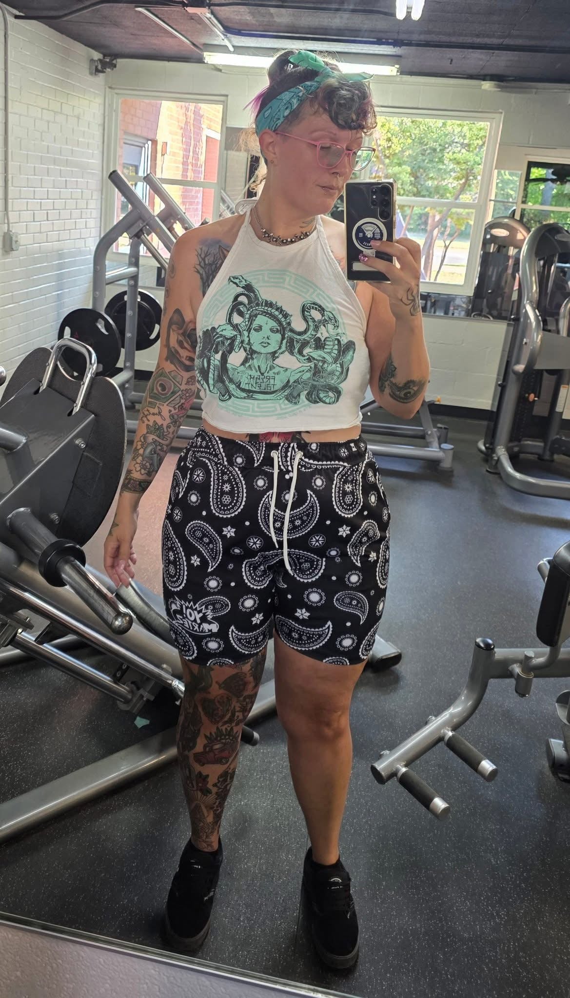 Katilyn Wink taking a mirror selfie in a gym, wearing a white halter top with a green graphic print and black paisley patterned Yo! Martez shorts in Firehouse Fitness Abilene Texas