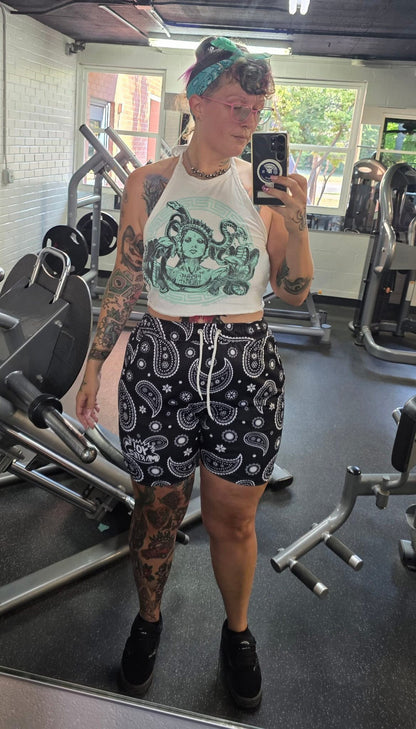 Katilyn Wink taking a mirror selfie in a gym, wearing a white halter top with a green graphic print and black paisley patterned Yo! Martez shorts in Firehouse Fitness Abilene Texas