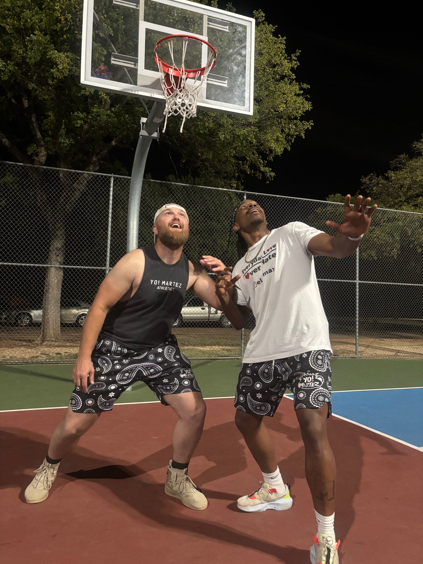 Anthony Hale aka Twon Hanks and Martez aka Yo! Martez playing basketball on an outdoor court at night under bright overhead lights. One man is wearing a white t-shirt and black paisley shorts, while the other wears a black tank top and matching black paisley shorts branded Yo! Martez