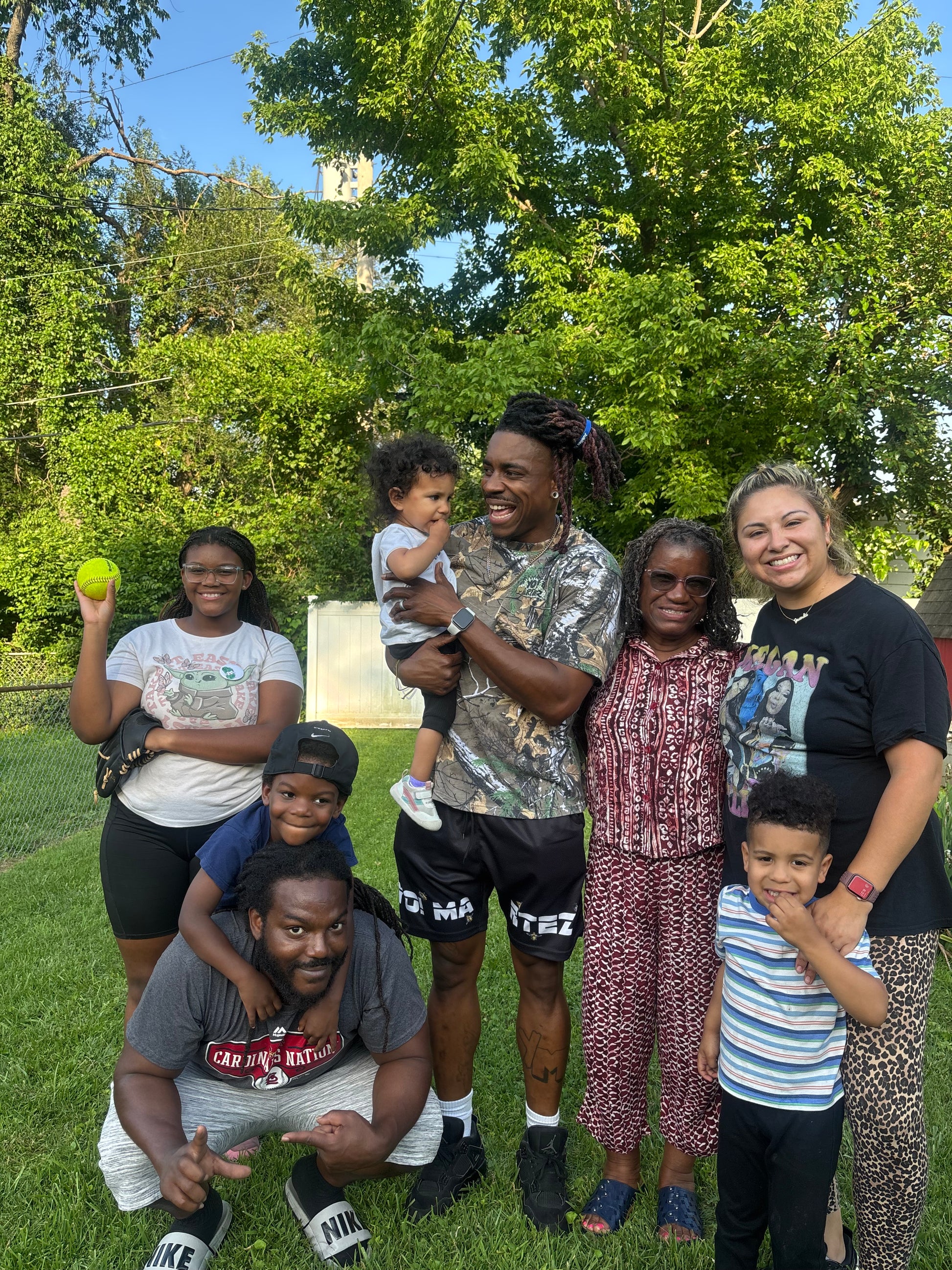 A group of seven people posing together outdoors on a grassy lawn under a large green tree. The group includes adults and children wearing casual clothing, with one person holding a yellow tennis ball.