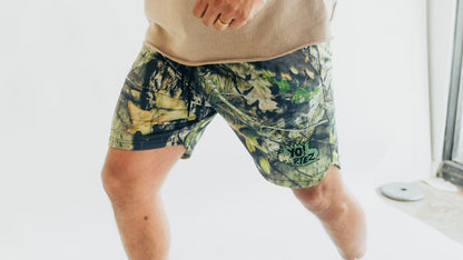 A person wearing camouflage print shorts with a green and black 'You Killz' logo patch, paired with a beige knit top, shown from the waist down in a bright indoor setting.