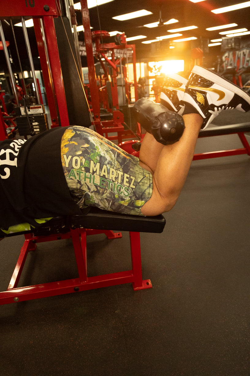 A person lying on a red gym bench performing a leg extension exercise, wearing camouflage-patterned athletic shorts with 'YOI MARTEZ ATHLETICS' printed on the side, a black t-shirt, and black-and-white sneakers in a gym setting.