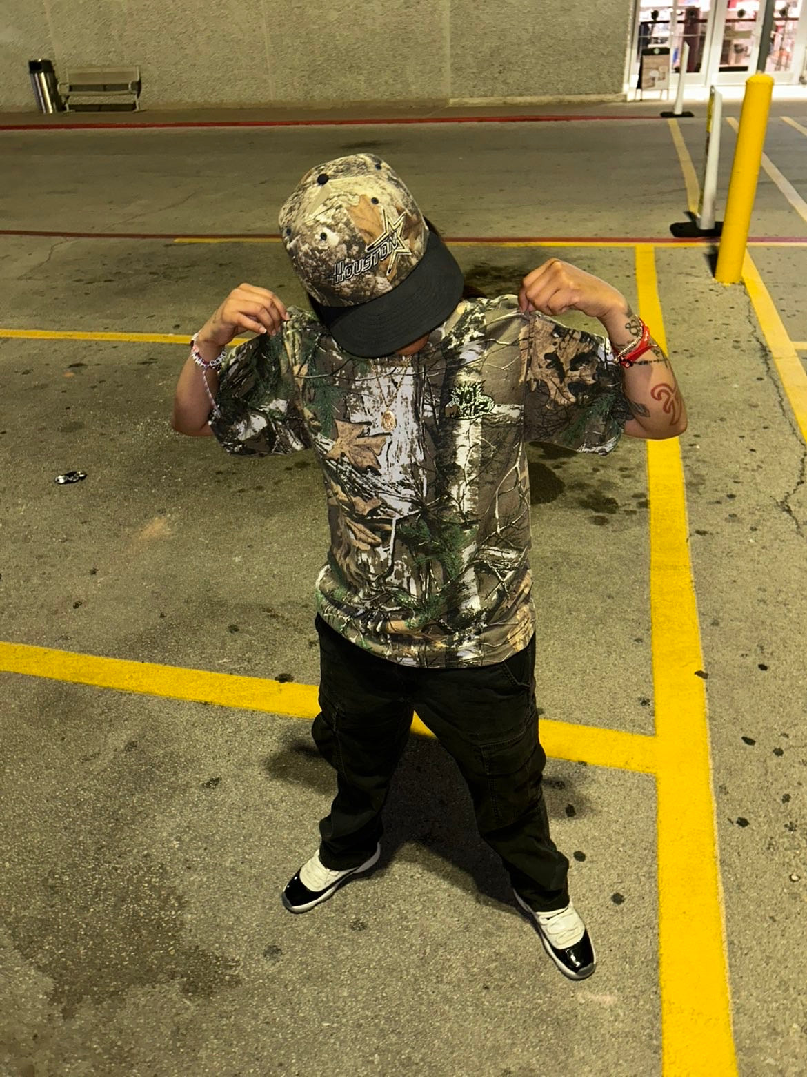 A person standing in a parking lot wearing a camouflage bucket hat with a black brim, a matching camouflage short-sleeve shirt, black pants, and black-and-white sneakers.