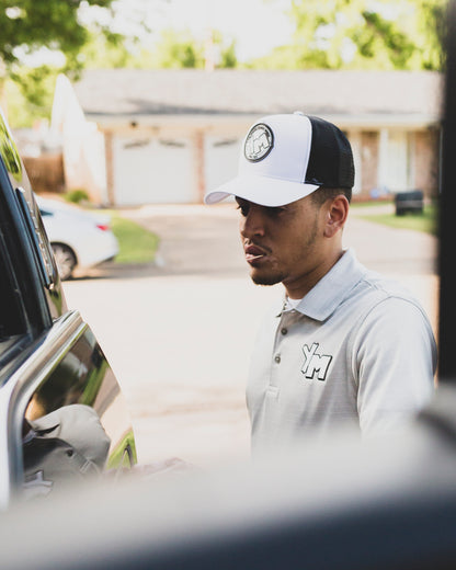 Jacob Cotton wearing a white and black Yo! Martez trucker hat and a light gray polo shirt with a stylized 'YM' logo on the chest, standing next to a car in a residential driveway. Photography by Rashad Barnes