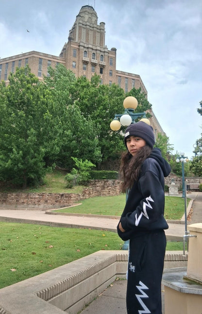 A person wearing a black beanie, black hoodie, and black sweatpants with white zigzag graphics stands outdoors in front of a large historic building and green trees.