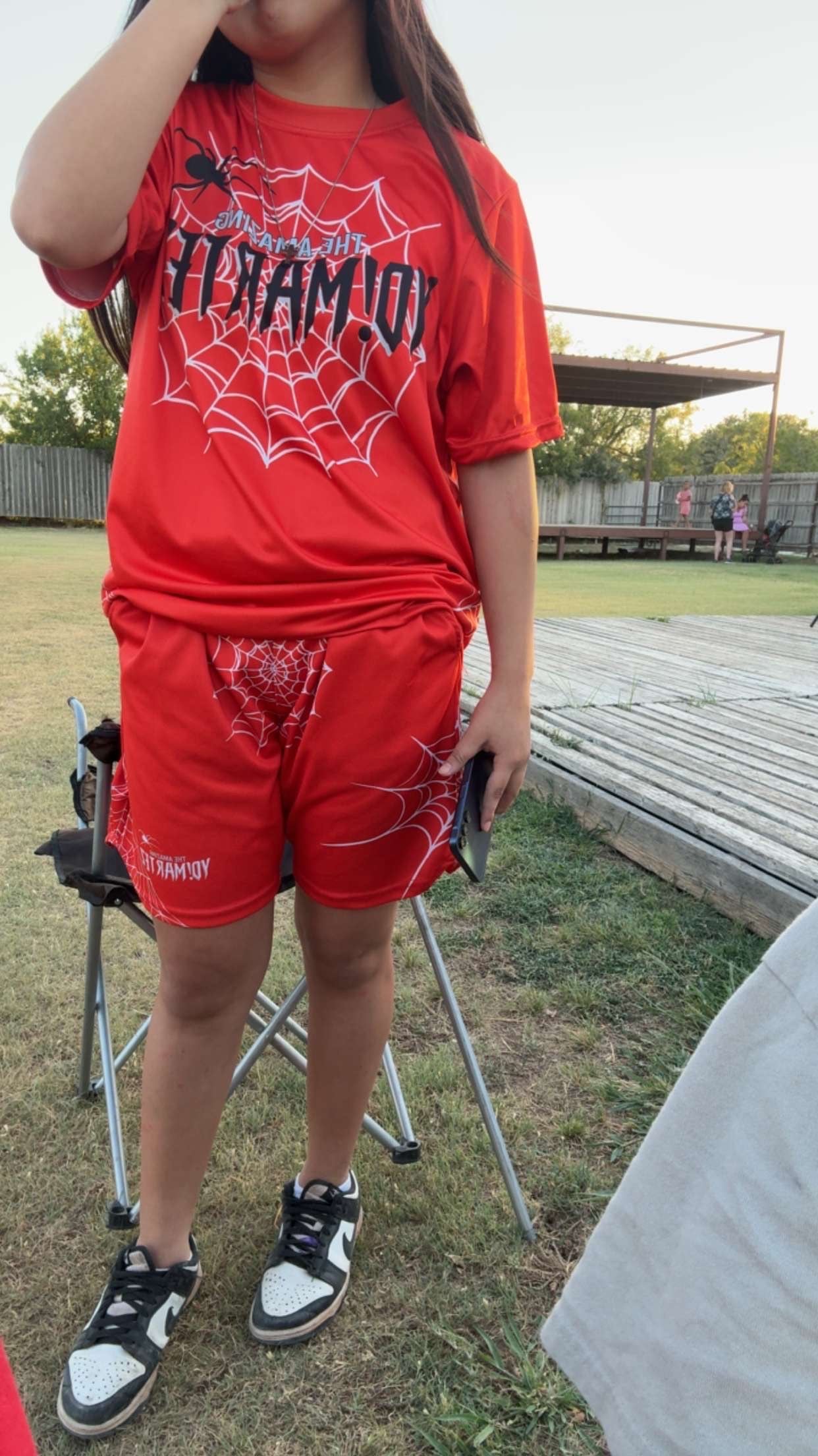 A person outdoors wearing a red short-sleeve T-shirt and matching red shorts with black and white spiderweb graphics and the word 'YO! MARTEZ' across the chest. The photo is taken in a grassy outdoor area with a folding chair and wooden structure in the background.
