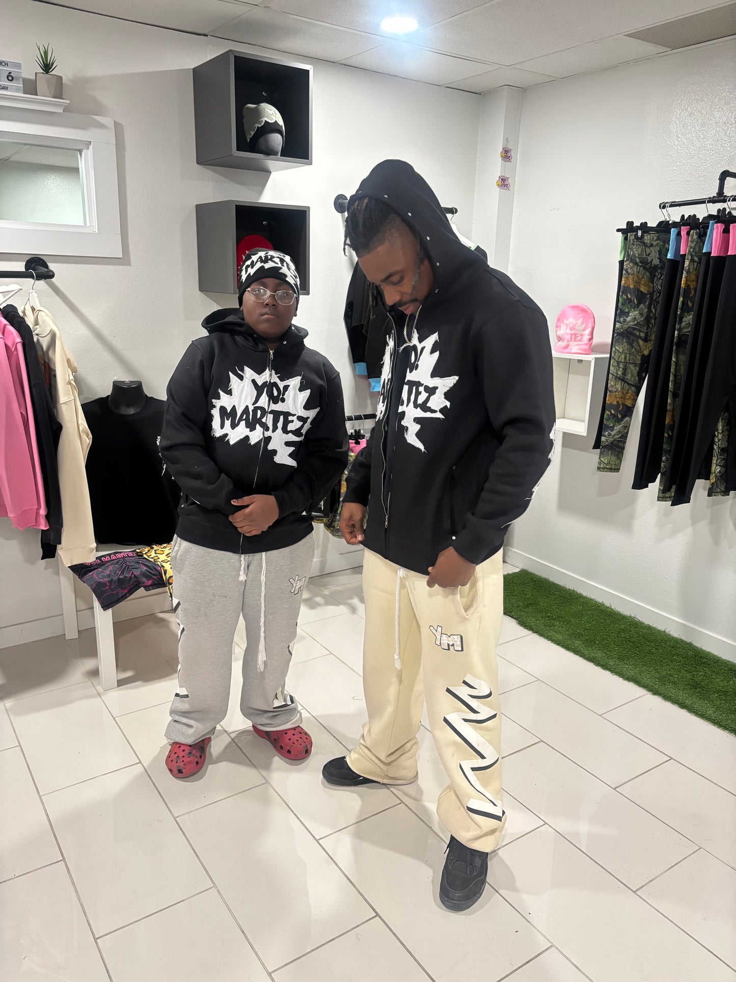 Martez & Braylon in Yo! Martez clothing store wearing black graphic hoodies with white 'Yo! Martez' text on the chest, paired with beige and gray pants and a black beanie respectively. The clothing store has white tiled floors, white walls, and clothing racks giving a luxuries aesthetic 